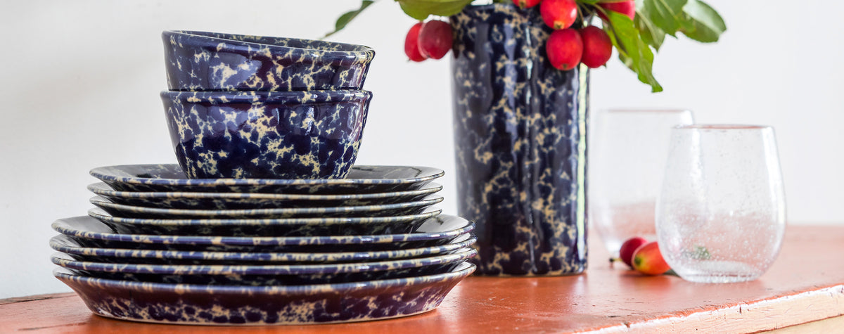 A stack of our classic dinnerware in customer favorite Blue Agate glaze.  A dark blue speckled on a tan base color, each piece hand glazed and no two alike. Lunch and Dinner plates shown along with medium basic bowls.  The tall utensil crock is in the background, doubling as a vase here with bright foliage for the season.